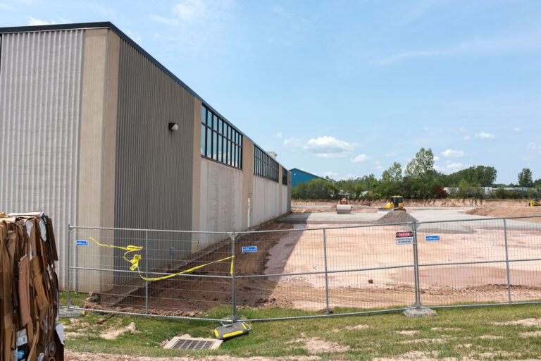 A fenced construction site with earthmoving equipment beside a Sealing Devices production building.