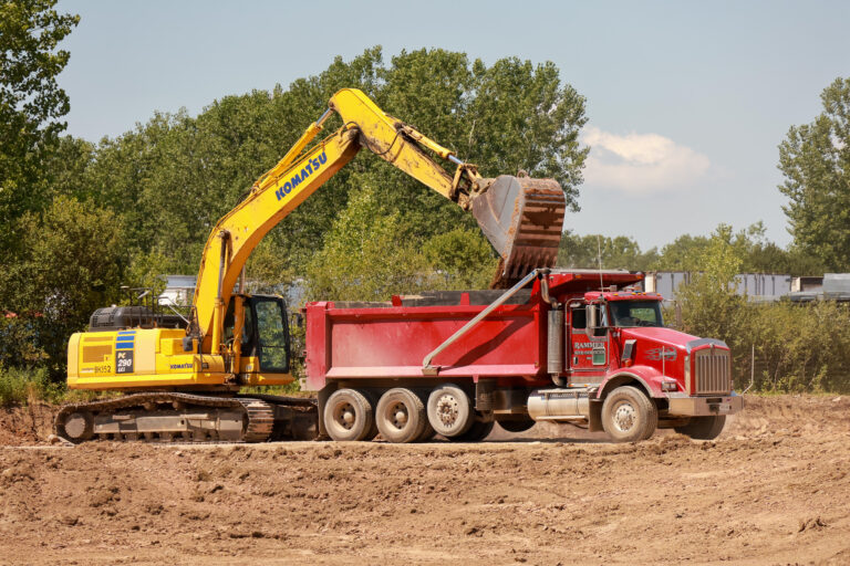 An excavator loads soil into a red dump truck at the Sealing Devices construction site.