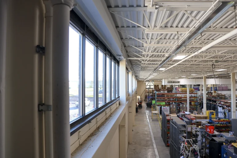 Elevated view of the manufacturing floor, with large windows, equipment, storage racks, and exposed ceiling trusses.