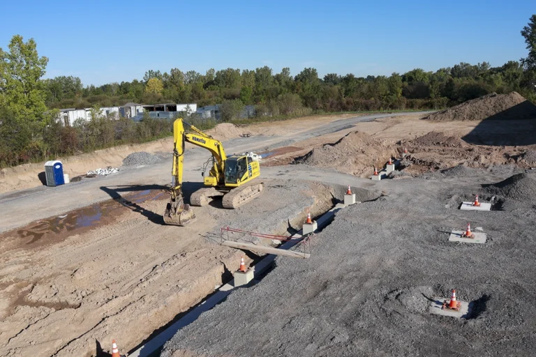 Yellow excavator beside foundation trenches and concrete footings marked with orange safety cones.