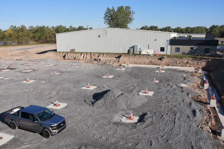 Elevated view of foundation pads, perimeter trenches, and a pickup truck beside the existing facility.