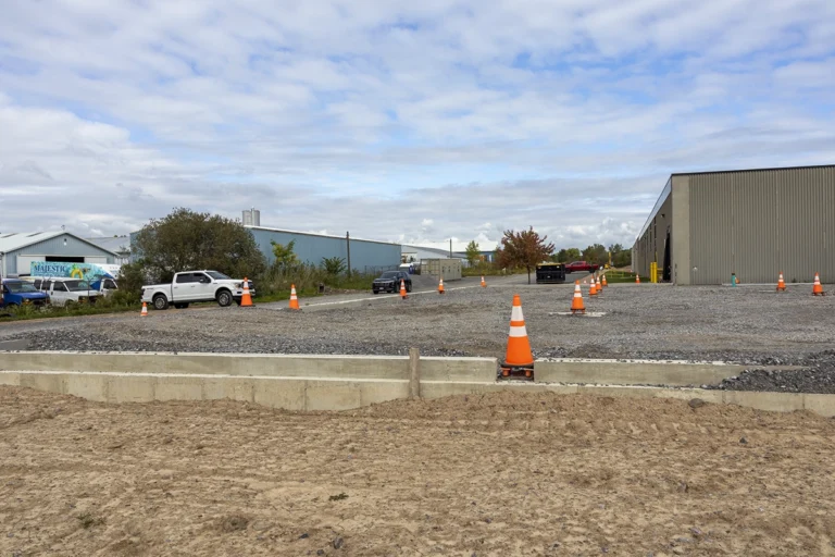 Concrete foundation walls bordering a gravel construction area marked with orange cones beside industrial buildings.