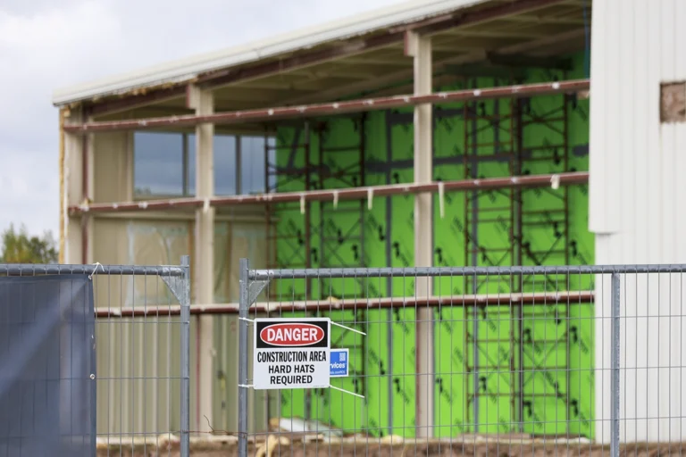 Construction fencing and hard-hat warning sign in front of exposed wall framing and green sheathing.